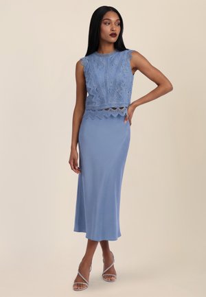 Frau trägt ein ärmelloses blaues Spitzen-Crop-Top und einen passenden Midi-Rock, steht mit einer Hand in der Hüfte und schaut zur Seite.