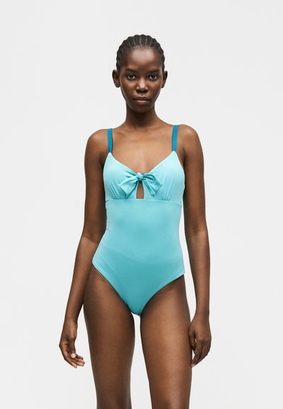 Γυναίκα με πλεξούδες που φοράει ολόσωμο μαγιό ανοιχτού γαλάζιου χρώματος με σκούρα μπλε τιράντες και λεπτομέρεια φιόγκου μπροστά, στεκόμενη μπροστά σε απλό φόντο.