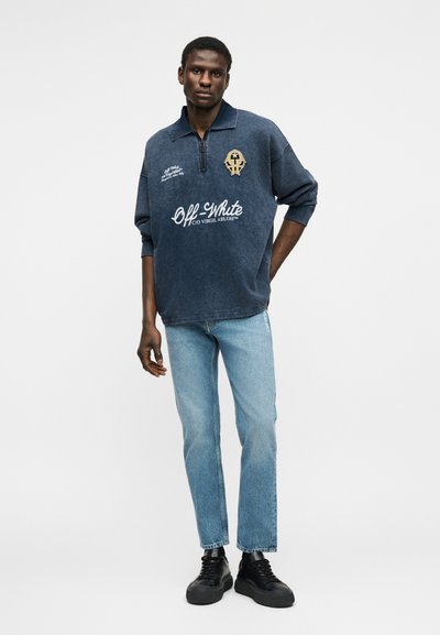 Ανδρικό μοντέλο που φοράει μπλε ναυτικό πουλόβερ Off-White με λευκό λογότυπο, ανοιχτόχρωμα μπλε τζιν και μαύρα αθλητικά παπούτσια, στέκεται με το ένα χέρι πίσω από την πλάτη.