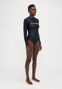 Čierny dlhý rukáv rash guard s bielym logom "BILLABONG" na prednej strane, spárovaný s čiernymi bikinami. Hladká textura, priliehajúci strih.