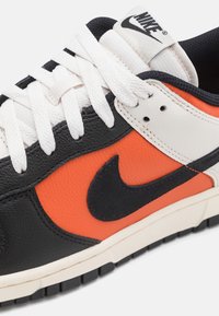 Ná мужчinné sneakers Nike z černé kůže, s výraznými oranžovými panely, bílými akcenty a texturovanými šňůrkami. Výrazné černé logo swoosh na boku.