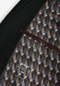 Doublure en tissu à motif géométrique à l'intérieur d'un vêtement noir avec bordure marron, étiquetée "Next Athletic Fit 40R Made in Vietnam."