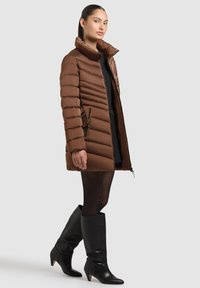 Abrigo de plumas marrón con diseño acolchado horizontal, cuello alto y bolsillos con cremallera. Modelo combinado con botas negras hasta la rodilla.