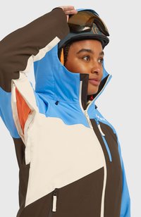 Hiihtotakki, jossa on väriblokkimuotoilu sinisessä, ruskeassa ja kermassa. Teksturoitu materiaali, jossa on vetoketjulliset taskut ja huppu. Sisältää verkkovuoren.