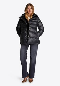 Schwarze Pufferjacke mit glänzender Oberfläche, ausgestattet mit einer Kapuze, frontalen Knöpfen und seitlichen Taschen. Getragen über einem schwarzen Oberteil und blauen Jeans.