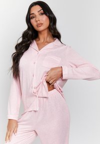 Lichtroze pyjamaset met een geribbelde textuur, een knoopsluiting bovenstuk met een zak en broek met elastische taille. Aangevuld met witte afwerking.