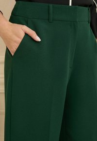 Pantaloni verdi su misura con una texture liscia, tasche laterali e un design frontale piatto con passanti per cintura.