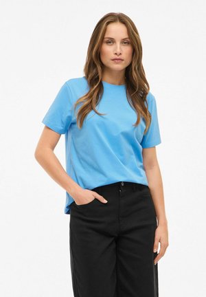 Jasnoniebieski t-shirt z krótkim rękawem wykonany z miękkiego materiału, z okrągłym dekoltem, zestawiony z czarnymi, szerokimi spodniami, ręce w kieszeniach.