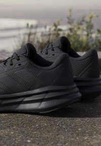 Paire de chaussures de course noires avec côtés texturés et semelles Cloudfoam placées sur un sol extérieur près de plantes.