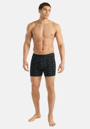 Athletischer Mann, der barfuß schwarze Boxershorts mit einem dezenten geometrischen Muster trägt und seitlich auf einem weißen Hintergrund schaut.