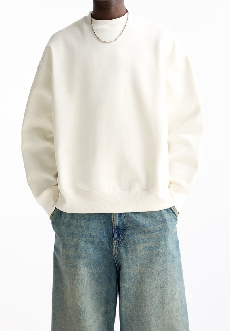 Personne portant un sweat-shirt uni blanc cassé et un jean bleu ample, les mains dans les poches, avec un collier en chaîne argenté.