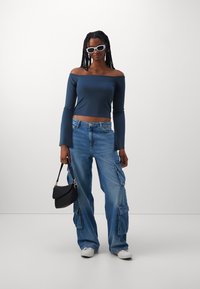 Mornarsko off-shoulder crop top s dugim flared rukavima, u kombinaciji s svijetloplavim cargo trapericama s bočnim džepovima i crnom torbom. Bijele sunčane naočale.