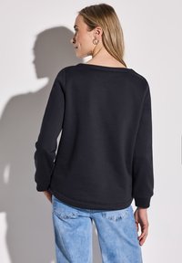 Sudadera negra de manga larga y escote amplio, combinada con jeans de un azul claro. La tela parece suave y tiene una textura lisa.