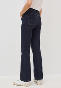 Jean évasé bleu foncé en denim, comportant deux poches arrière, des coutures visibles et une taille haute. À porter avec des baskets blanches.