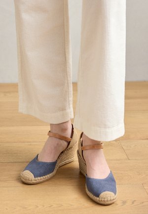 Mėlyni drobės platforminiai sandalai su nupinta jute baze, turintys kulkšnies dirželį ir tekstūruotą kreminės spalvos kojų dangtelį. Dėvimi su kreminės spalvos kordūro drabužiais.