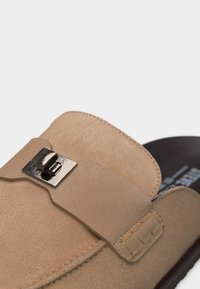 Κοντινό πλάνο σε μια μπεζ σουέτ γόβα χωρίς κορδόνια με ασημί ορθογώνια αγκράφα στον πάνω ιμάντα και μαύρη εσωτερική σόλα.