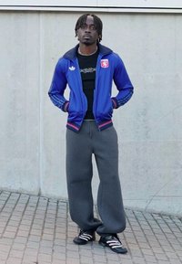 Blue athletic jacket with red accents, black shirt, gray sweatpants, and black shoes with white stripes. Standing against a concrete background.