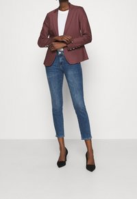 Burgundowa marynarka z wąskimi klapami i srebrnymi guzikami, w zestawieniu z niebieskimi jeansami do kostek i czarnymi szpilkami.