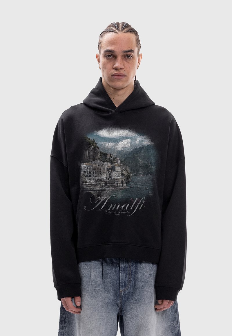 Personne portant un sweat à capuche noir avec imprimé pittoresque de la côte amalfitaine et texte, associé à un jean bleu délavé ample, debout devant un fond uni.