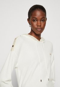 Hoodie bianco con tessuto testurizzato, cappuccio beige chiaro a contrasto e dettagli con bottoni in tono oro sulle spalle. Presenta lacci con punte metalliche.