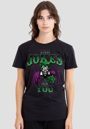 Young woman with long brown hair wears a black t-shirt showing a green-haired Joker in a purple suit and text "The Joke's on You."