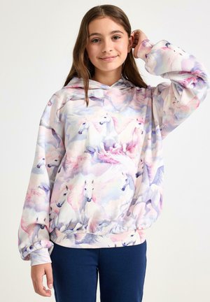 Pitkähiuksinen tyttö, jolla on päällään pastellinsävyinen huppari, jossa on valkoisia hevosia ja vaaleanpunaisia pilviä, seisoo yksinkertaisen valkoisen taustan edessä.