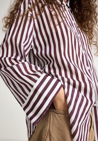 Pruhovaná košile v barvách mahagonové a bílé, vyrobená z lehké látky. Rukávy jsou široké s knoflíkovým zapínáním. Viditelná kapsa.