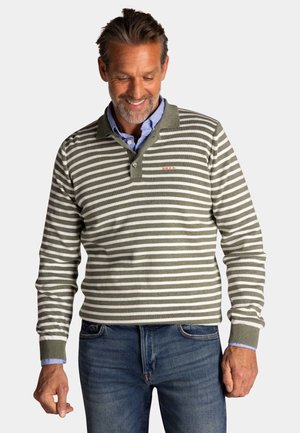 Man die een groen-wit gestreept poloshirt met lange mouwen draagt over een blauw overhemd met knoopjes en een blauwe spijkerbroek, glimlachen en naar beneden kijkend.