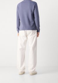 Person som står med ryggen vänd, iförd en lila långärmad tröja, vita lösa byxor och vita sneakers framför en enfärgad bakgrund.