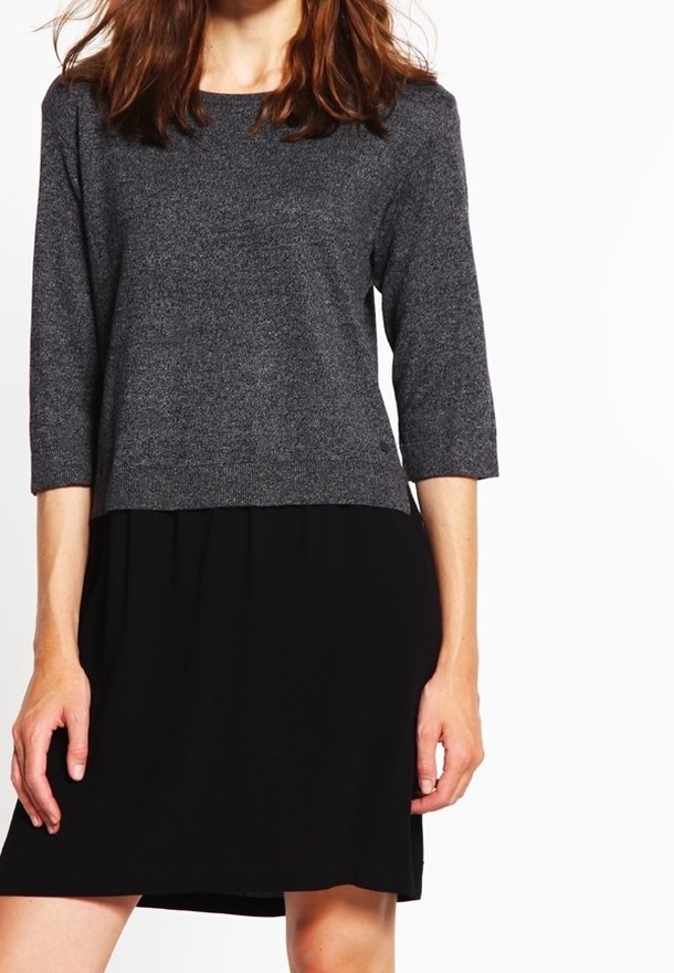 Femme portant un pull gris foncé à manches trois-quarts sur une jupe noire longueur genoux, debout devant un fond blanc.