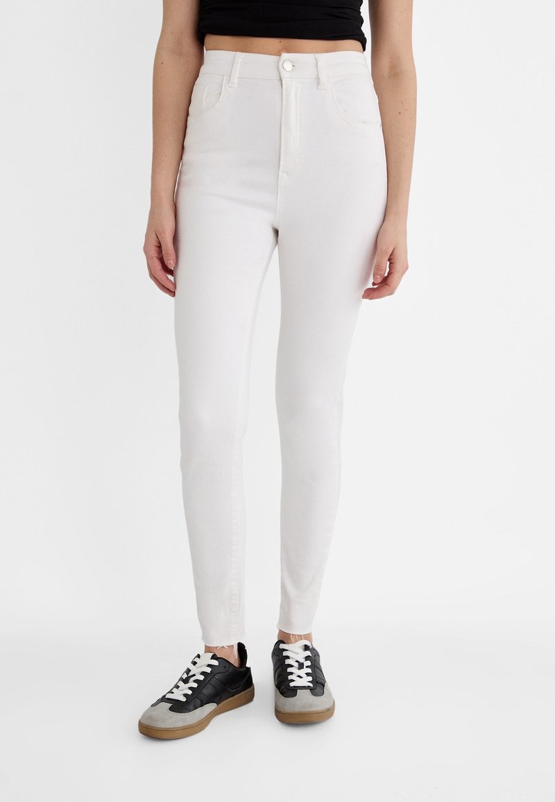 Jean skinny taille haute blanc en denim, avec cinq poches, une fermeture à bouton à l'avant et une texture lisse. Associé à des baskets noires et grises.