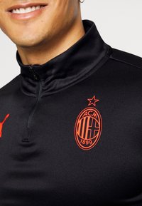 Giacca nera con zip realizzata in tessuto morbido, caratterizzata da un logo Puma rosso e dall'emblema dell'AC Milan con una stella sopra, evidenziando l'identità della squadra.