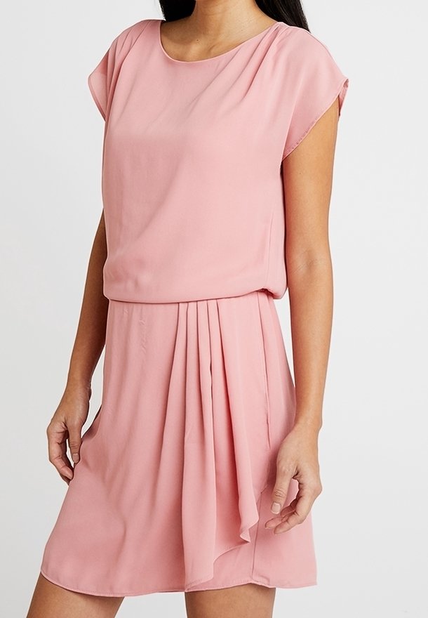 Femme portant une robe rose clair à manches courtes avec un détail plissé sur la jupe et un col rond sur un fond uni.