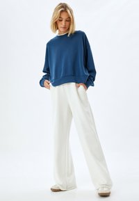 Felpa blu in pile con scollatura rotonda, vestibilità comoda e orlo a coste, abbinata a pantaloni bianchi a gamba larga e sneakers beige.