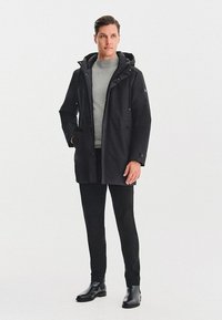 Czarna wodoodporna kurtka z kapturem, o eleganckim fasonie, z przednimi kieszeniami i stonowanym przeszyciem. Nosi ją osoba w szarym swetrze i czarnych spodniach.