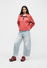 Γυναίκα που στέκεται με τα χέρια πίσω από την πλάτη, φορώντας κοραλλί fleece πουλόβερ, ανοιχτό μπλε παντελόνι φαρδύ με σκισίματα και κόκκινα αθλητικά παπούτσια σε λευκό φόντο.