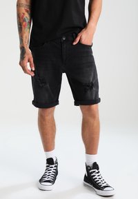 Pantalones cortos de mezclilla negra con puños enrollados, detalles desgastados en la parte frontal y un ajuste ajustado. Combinados con zapatillas altas negras y calcetines blancos.