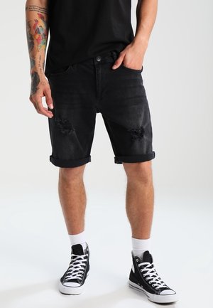 Pantalones cortos de mezclilla negra con puños enrollados, detalles desgastados en la parte frontal y un ajuste ajustado. Combinados con zapatillas altas negras y calcetines blancos.