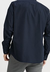 Granatowa koszula z długim rękawem o gładkiej fakturze, z mankietami na guziki, prostym dołem i detalem na karczku z tyłu. Prosty, dopasowany krój.