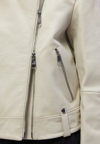 Κρεμ δερμάτινο μπουφάν με εφαρμοστή γραμ μή, ασύμμετρο μπροστινό φερμουάρ και πολλαπλές τσέπες με φερμουάρ, διακοσμημένες με ασημένιες λεπτομέρειες.