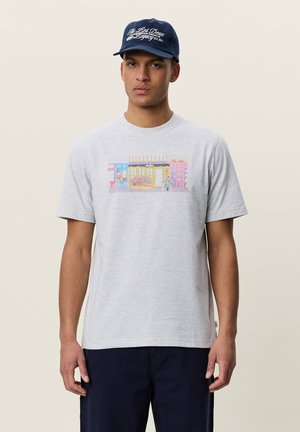 Jeune homme portant un t-shirt gris clair avec une impression colorée d'une vitrine de magasin et une casquette bleu foncé avec un texte blanc.