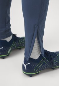 Pantaloni sportivi blu con lati con cerniera, abbinati a scarpe da ginnastica blu scuro con dettagli verdi e superfici texturizzate. Calze grigie visibili sopra le scarpe.