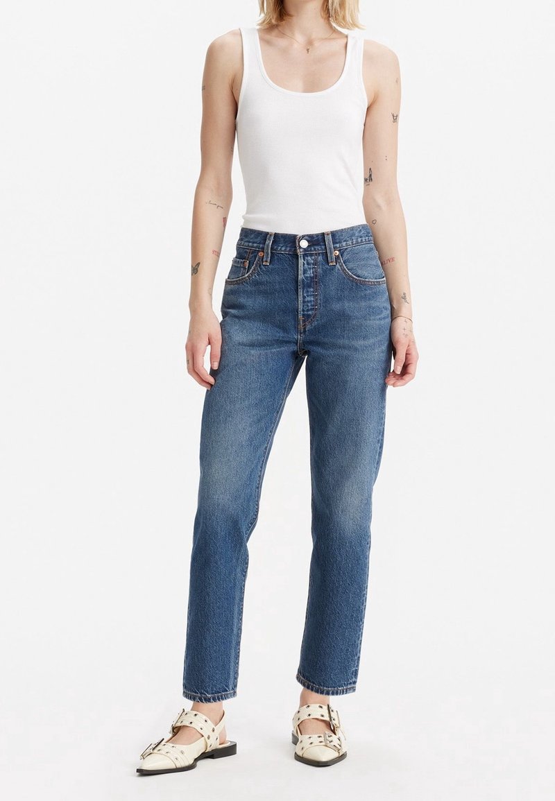 Personne debout portant un débardeur blanc, un jean bleu taille haute et des sandales blanches à clous sur un fond blanc uni.