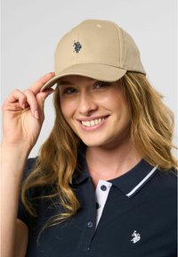 Beige baseball cap with a curved brim, featuring navy blue embroidered logo at the front. Worn by a person with shoulder-length hair.