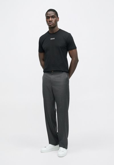 Μαύρο βαμβακερό μπλουζάκι με το λογότυπο "VERSACE", συνδυασμένο με γκρι ραμμένο παντελόνι και λευκά αθλητικά παπούτσια. Το μοντέλο στέκεται με τα χέρια σταυρωμένα.