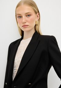 Blazer nero con un design strutturato e accessori in oro, indossato sopra un top in pizzo bianco, che mette in mostra intricate trame e texture.