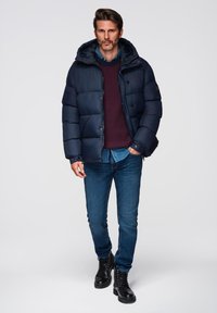 Granatowa kurtka pufowa z odpinanym kapturem, zapięciem na guziki oraz pikowaną fakturą, noszona na burgundowym swetrze dzianinowym i niebieskich dżinsach. Czarne buty lace-up.