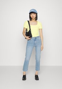 Top amarelo de mangas curtas com nervuras, jeans de perna reta azul claro, mules pretos de salto, e um boné de denim com "Levi's" bordado na frente.