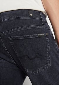 Jeans de mezclilla azul oscuro con un bolsillo trasero que presenta un patrón de olas, con una pequeña etiqueta negra y acentos de botón plateado.