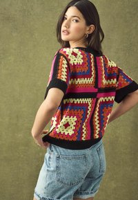 Top kratkih rukava u heklanom stilu s višebojnim uzorcima u crvenoj, narančastoj, žutoj, plavoj i crnoj boji, uparena s traper kratkim hlačama.